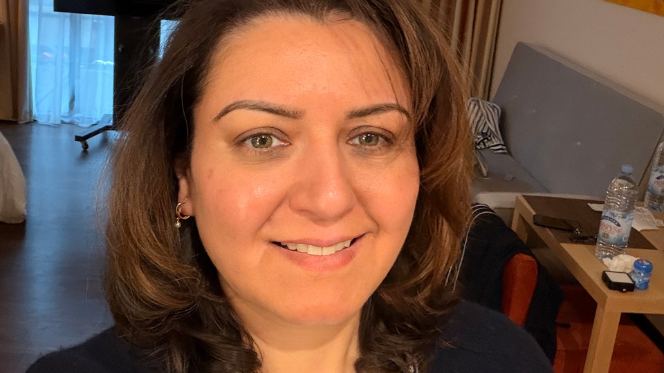Raina takes a selfie, smiling for the camera. She has shoulder-length brown hair which curls around her chin and forehead. She wears a black cardigan over a white tshirt