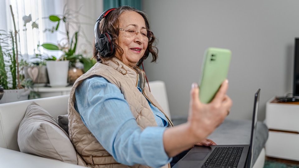 Woman sits on a sofa with a laptop, holding a phone out in front of her