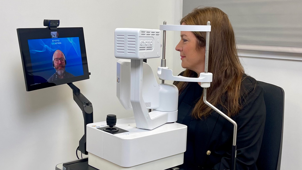 A woman is seated at a slit lamp. On a screen attached to the device is a video call with a remote clinician