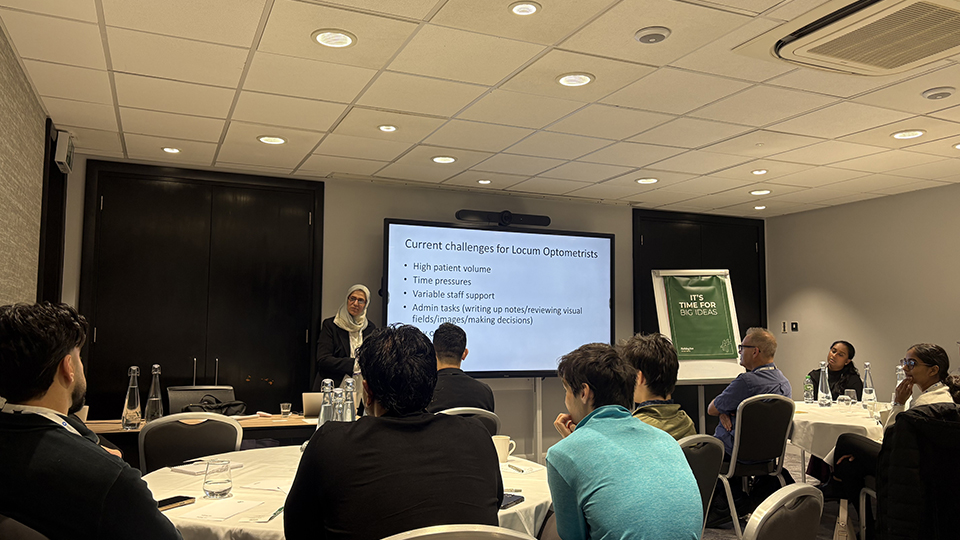 A woman wearing a light-coloured headscarf presents a slide titled “Current challenges for Locum Optometrists” to an audience seated around tables in a conference room