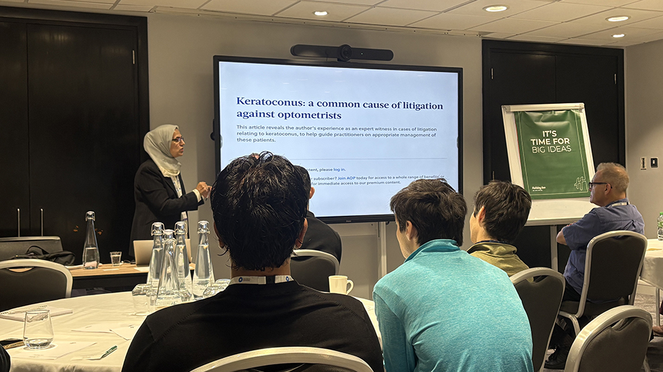 A woman wearing a light-coloured headscarf presents an article titled “Keratoconus: a common cause of litigation against optometrists” to an audience at a professional meeting. Attendees sit facing the screen, with a green “It’s time for big ideas” banner beside it
