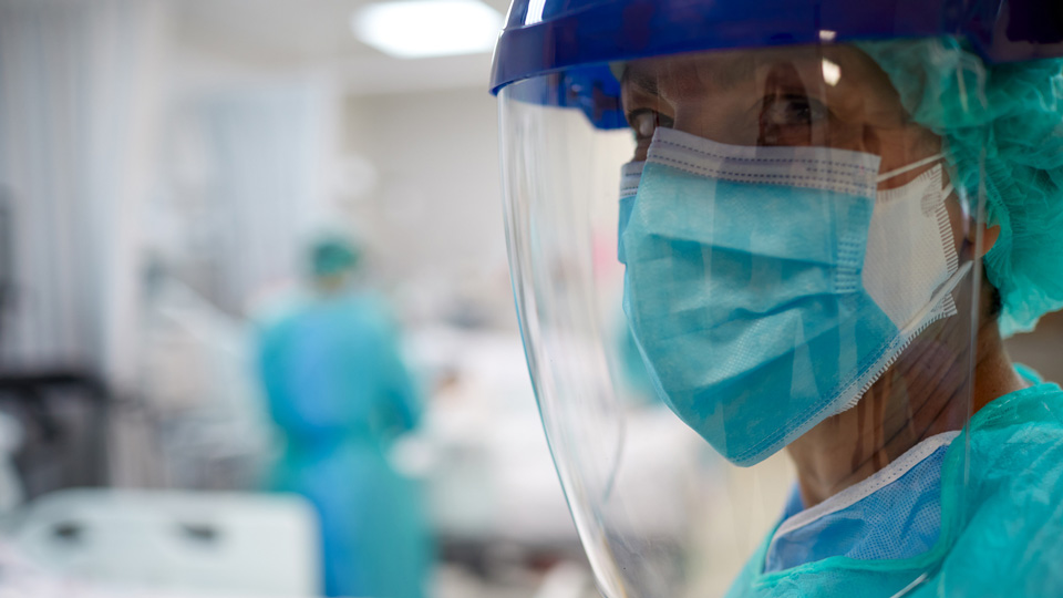 A clinician wearing a surgical mask and face shield stands in a hospital