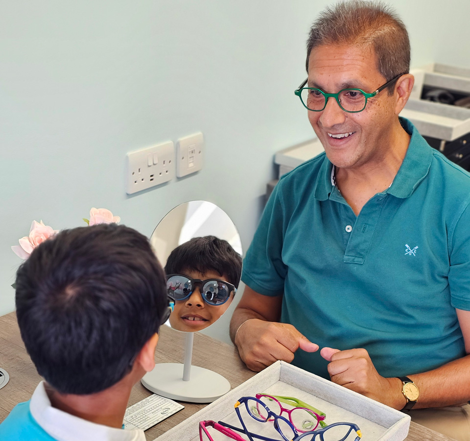 Harsh Shah is seated at a dispensing desk with a child who is trying on colourful pairs of sunglasses
