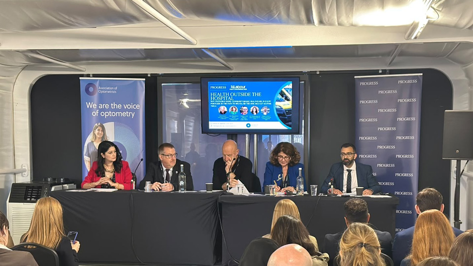 Five panellists sit behind a table at an Association of Optometrists event, taking part in a discussion on “Health Outside the Hospital,” with an audience seated in front of them. A presentation screen and banners for the Association of Optometrists and Progress stand behind the panel