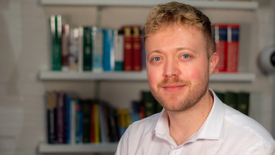 Portrait of a man with short, curly light brown hair and a beard, wearing a white collared shirt and looking towards the camera. Bookshelves filled with colourful books are visible in the background