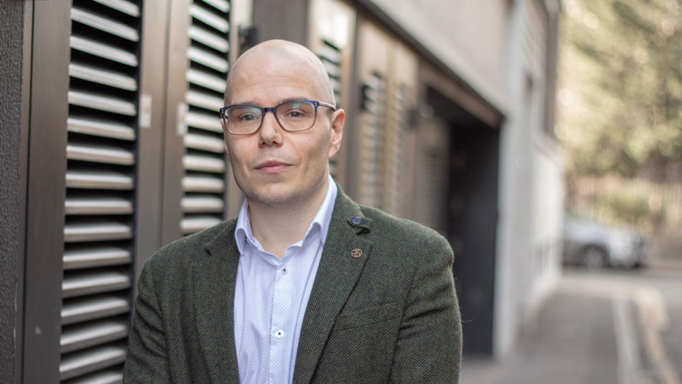 Portrait of a bald man wearing dark-framed glasses, a green tweed blazer with a lapel pin and a pale blue patterned shirt. He stands beside a modern building, with a pavement, parked cars and trees softly blurred in the background