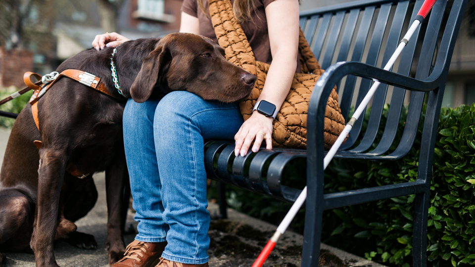 A woman sits on a park bench with a white cane and a service dog