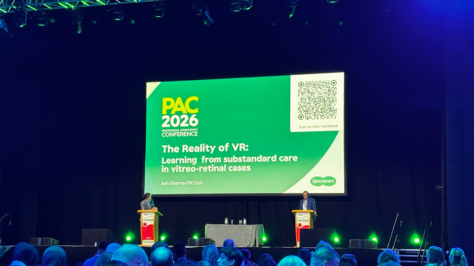 Two men stand at lecterns on a stage. Behind them is a large screen in green and white with the PAC 2026 conference logo and a title for the session: The reality of VR – learning from substandard care in vitreoretinal cases