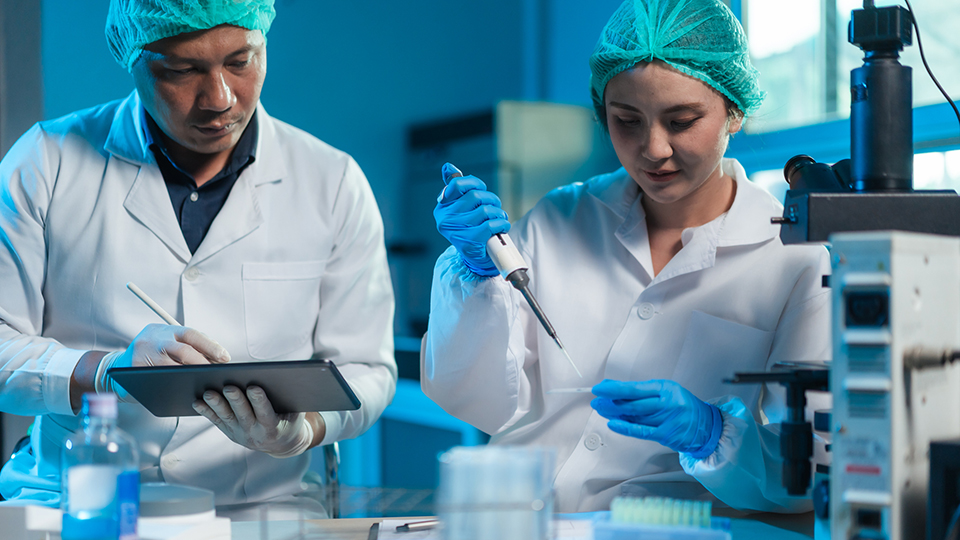 Two scientists work in a laboratory. One holds a clipboard while the other holds a pipette over test tubes