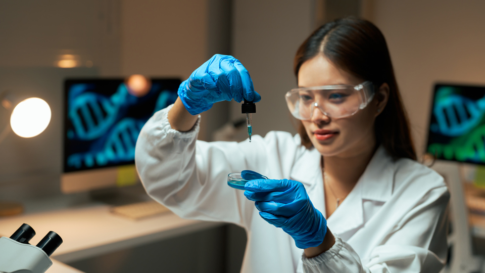 A scientist uses a pipette over a petri dish with a DNA double helix on a screen in the background
