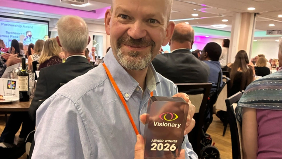 A man is seated at a circle event table dressed in a shirt and smart trousters. He holds a glass award for the Visionary awards