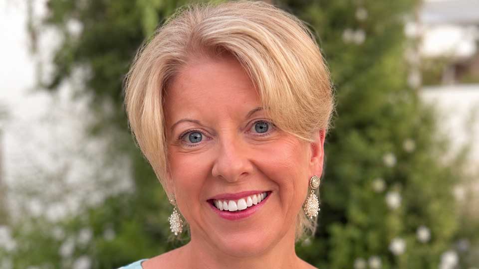 A smiley blonde woman in a blue floral dress and dangling pearl earrings