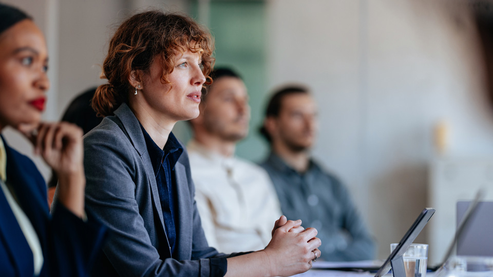 A woman listens among a group of people sitting at a board table