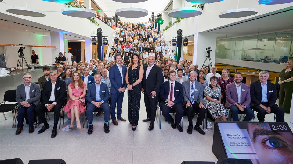 A large group of people pose for a photograph inside a modern venue, with three people standing at the front and others seated behind them on tiered steps. Lighting equipment and cameras surround the group, while a screen in the foreground displays “Welcome to The Vision Centre”