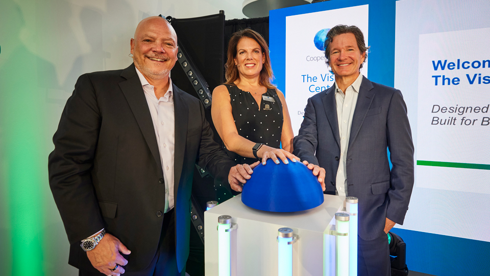 Three people pose beside a blue dome-shaped display at a CooperVision event, with their hands resting on the display and a presentation screen visible behind them