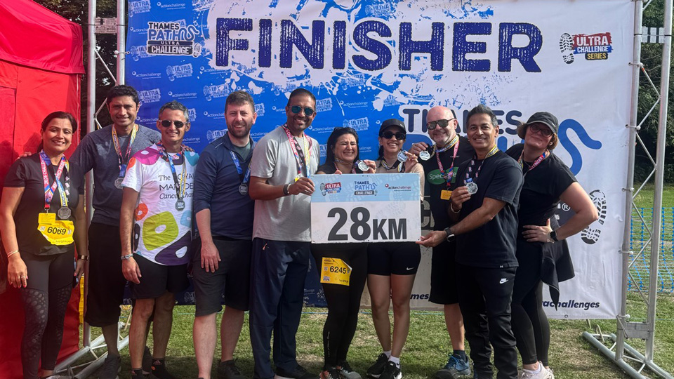 Ten people pose together at the finish line of the Thames Path Ultra Challenge, wearing medals and holding a sign marked “28KM”, beneath a large “FINISHER” banner. Several wear Specsavers-branded clothing, with a red event tent and trees in the background