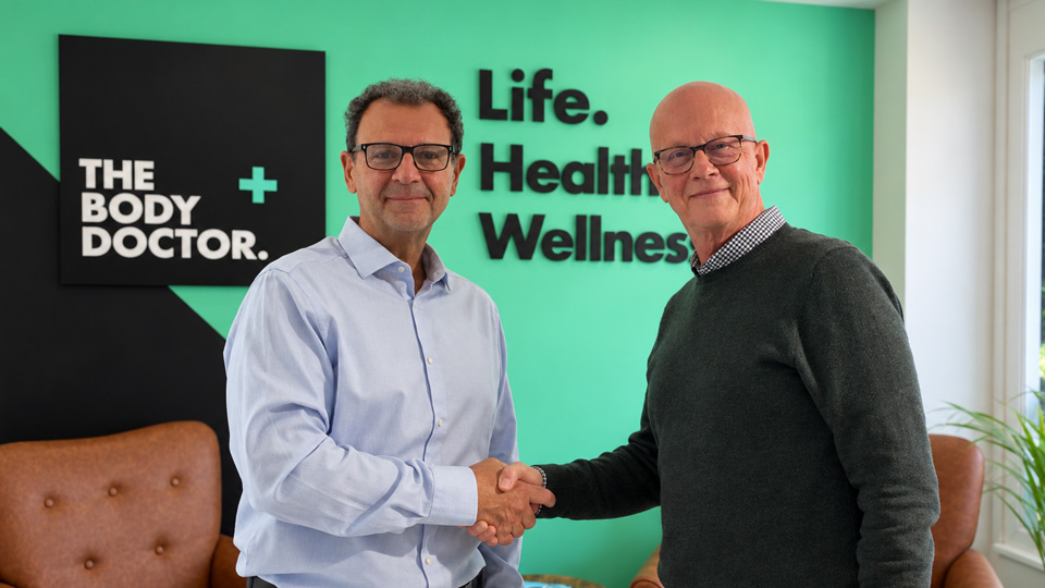 Two men in smart attire shake hands, smiling. The room is green and black with The Body Doctor logo