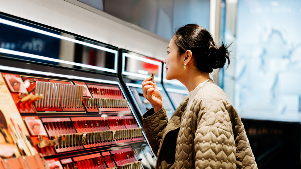 A woman tries on lipstick in front of a display of different cosmetic items