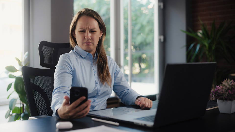 A woman in her 40s at a desk holds her smartphone away from her and squints