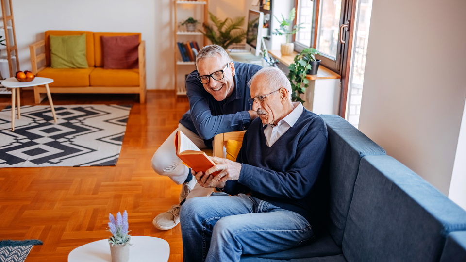 Middle age man reading book with his father while sitting close to window in living room