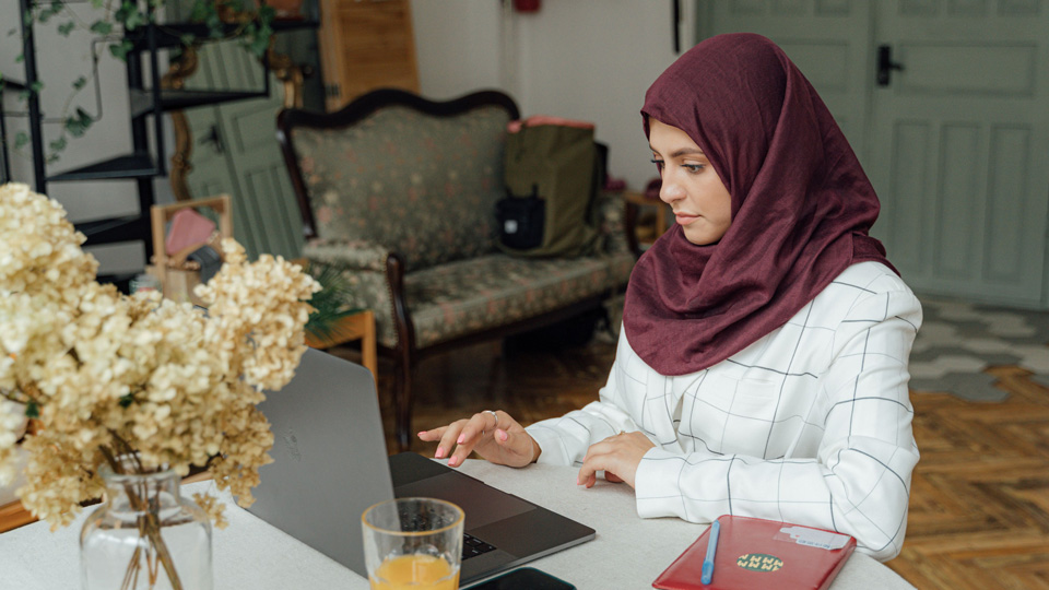 A woman is seated at a round table working on a laptop, she has a maroon headscarf and is surrounded by tools to make notes