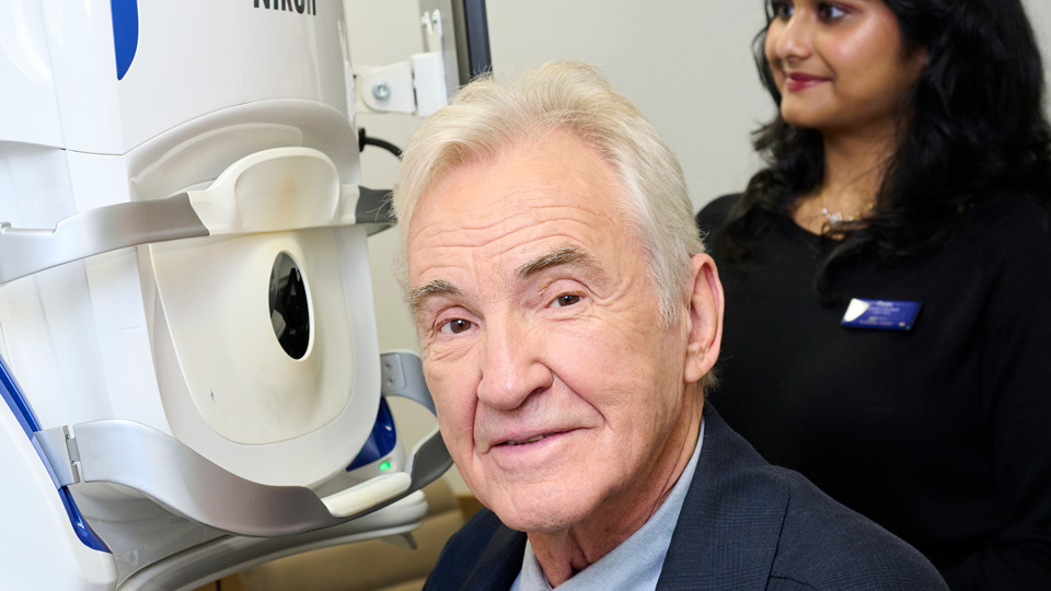 Larry Lamb is seated at a Nikon device as part of an eye test, though he has turned to look at the camera. Behind him, an optometrist looks at a screen