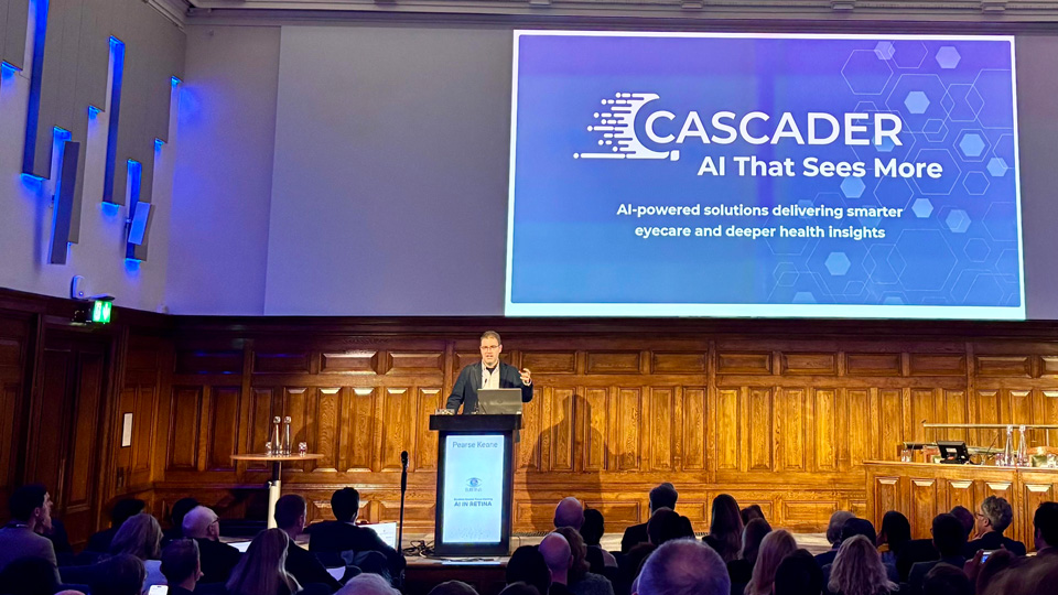 Professor Pearse Keane stands on stage behind a lectern in a large, grand room. A presentation holds the Cascader logo and information about AI solutions for smarter eye care