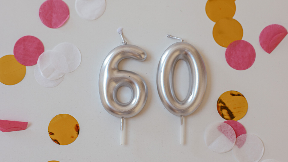 Arranged on a white background are two silver number candles spelling out 60 and a scattering of amber, white and pink confetti