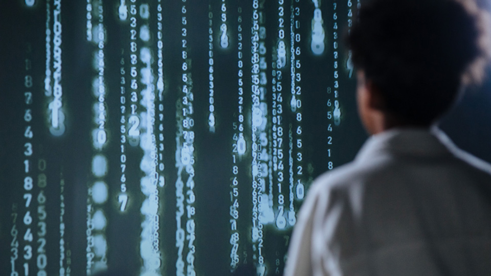A man with his back to the camera looks into a display of glowing numbers that drop down in lines from above, indicating data and artificial intelligence