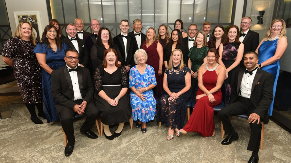 A group of people in black tie attire are gathered together for a celebratory picture. At the front and centre of the group is Dame Mary Perkins wearing an abstract blue and pink dress.