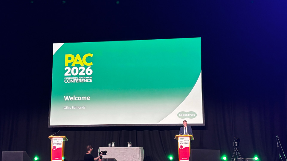 Giles Edmonds stands at a lectern on a large stage. Behind him is a welcome presentation for PAC 2026