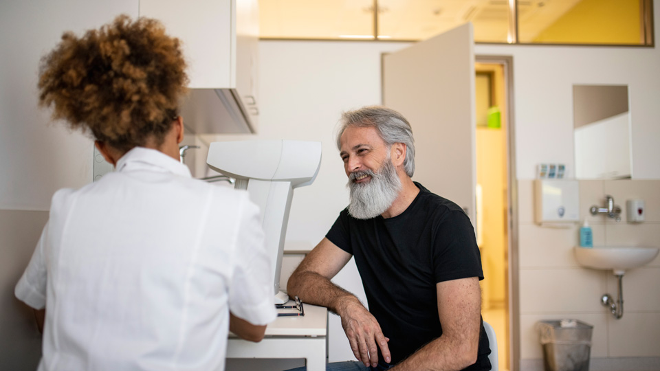 A patient with greying hair and beard speaks to a practitioner who is wearing a white clinical coat. They are seated in a consulting room