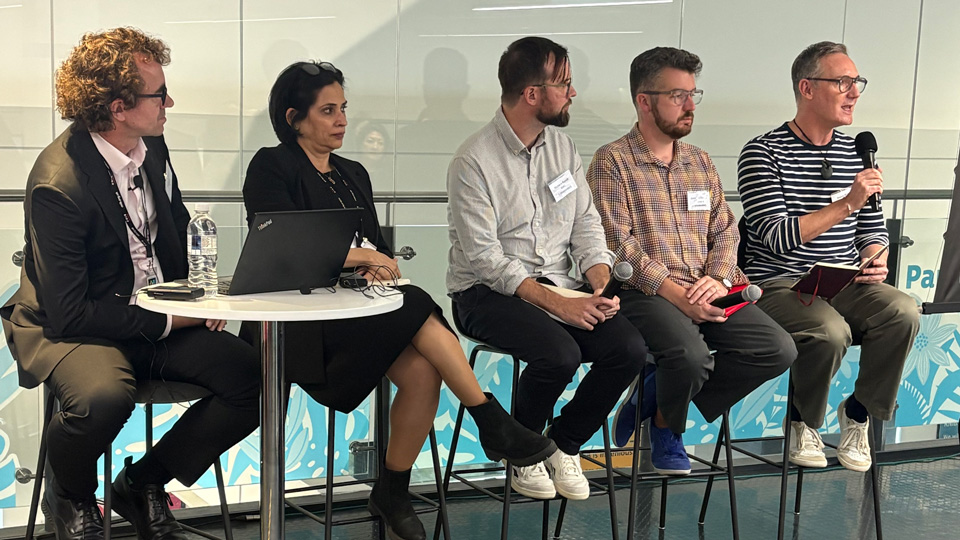 The Audio is King panel sitting on tall chairs next to one another in a line, in front of a large glass window in an office. From left to right they are John Paton and Sonali Rai from RNIB, Richard Kurzik from BBC, Jonathan Penny from Channel 4 and Simon Elder from Sky