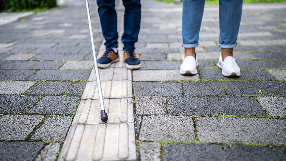 Legs and feel of two people, one with a white stick, walking along a pavement