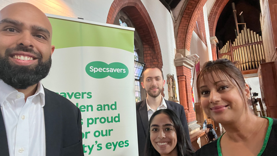 Two men and two women dressed in Specsavers uniform stand around a banner for eye care services