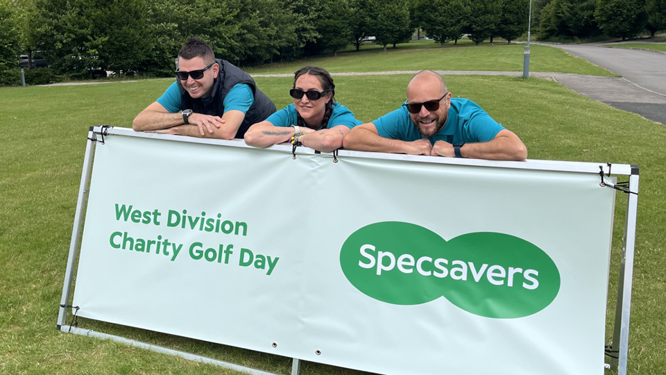 Three people pose with their heads popping up behind a sign that celebrates the Specsavers West Division charity golf day