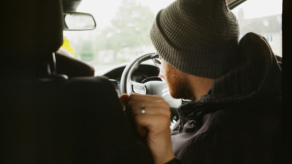 A photograph from the back seat of a car showing a driver wearing glasses and a knitted hat