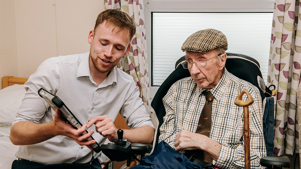 Sam is sat inside a house showing an elderly man the results of an eye test via an iPad