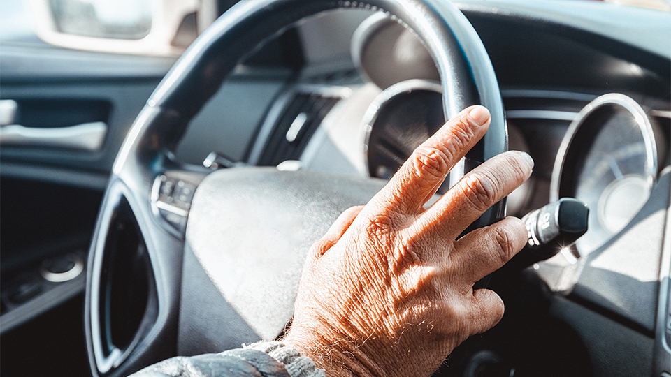 The hands of an older driver are shown steering the wheel of a car