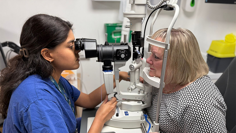 Roshni is wearing blue scrubs and is sat down, performing an eye test on a blonde female patient 