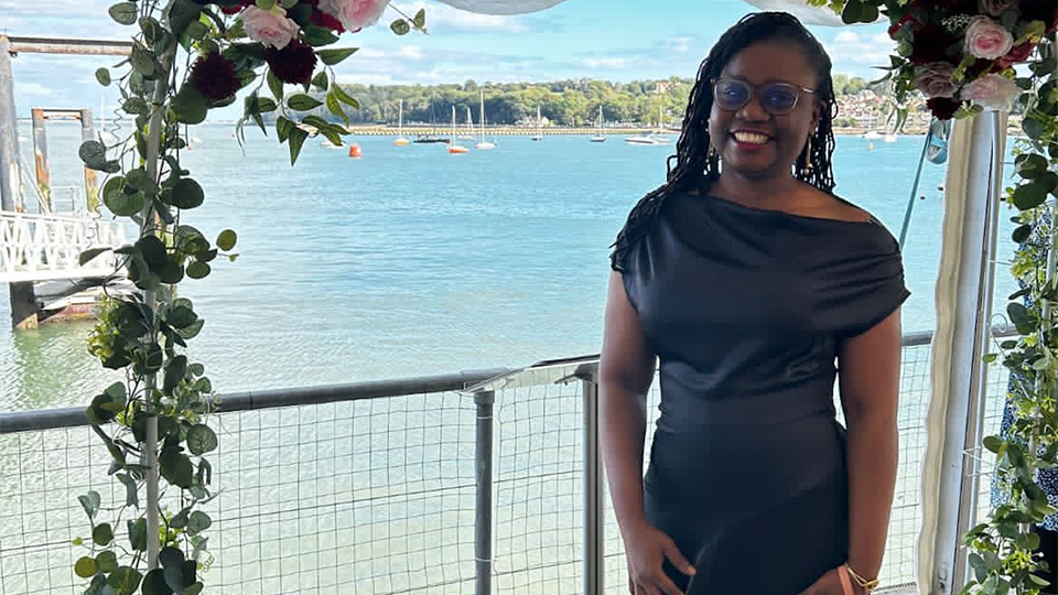 Chibogu wears a formal black dress with bright pink sandal heels. She stands beneath a white steel archway decked in trailing green vines and red and pink roses. Behind her is the beautiful crystal ocean and boats in a bay