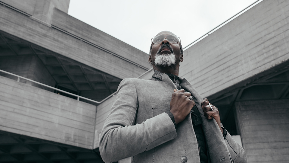 Antony wears a fine metal pair of glasses and wears grey, holding the lapels of his jacket and looking pensively into the grey sky 
