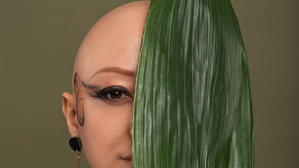 Morena poses behind a large green palm leaf. She wears heavy eye makeup in black and brown colours that wraps around her cheek and long earings with a circle in a black and brown marble design