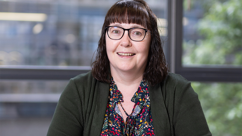 Professional head-and-shoulders portrait of a smiling woman with shoulder-length brown hair and fringe, wearing black rectangular glasses. She faces the camera whilst sitting at a table