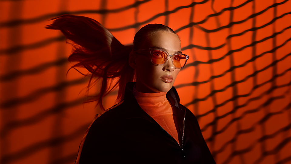 A woman in a ponytail and orange tinted spectacles stands in front of an orange background – dark shadows are cast in a net over her 
