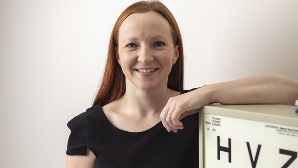Claire has long red hair and is wearing a black top, stood smiling with her arm resting on an Amsler chart 