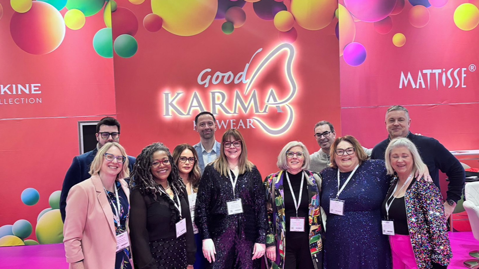 > The Good Karma team gather for a group photograph on the brand’s exhibition stand at 100% Optical. The stand is a coral-orange and colourful. Karma, lit up, has the wing of a butterfly integrated into the logo