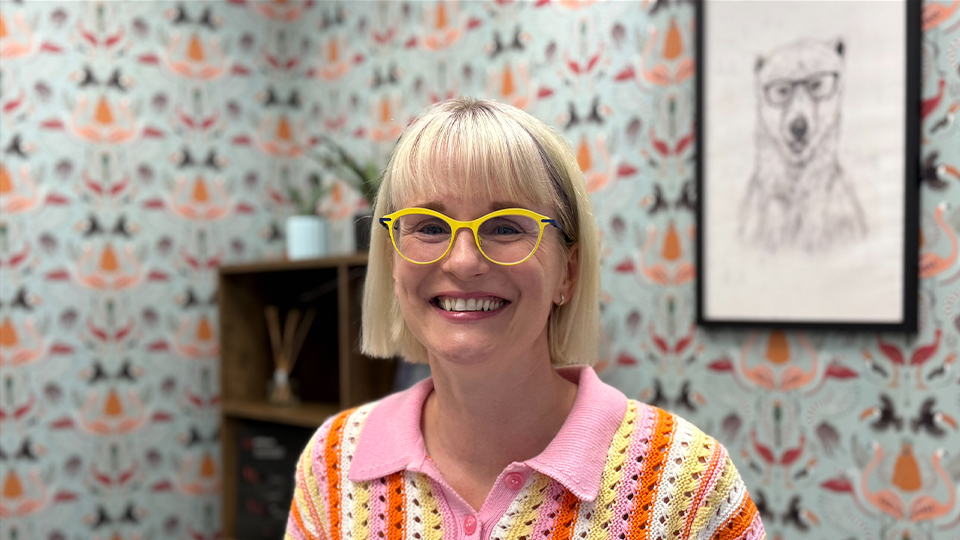Kirsty wears a pair of wired cat’s eye glasses in neon pink and beige with a colourful pink scarf in her hair