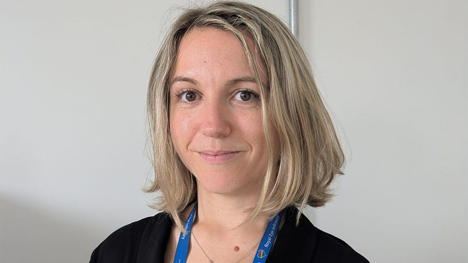 Liz has blonde hair and is stood against a white wall wearing a black top and blue lanyard 