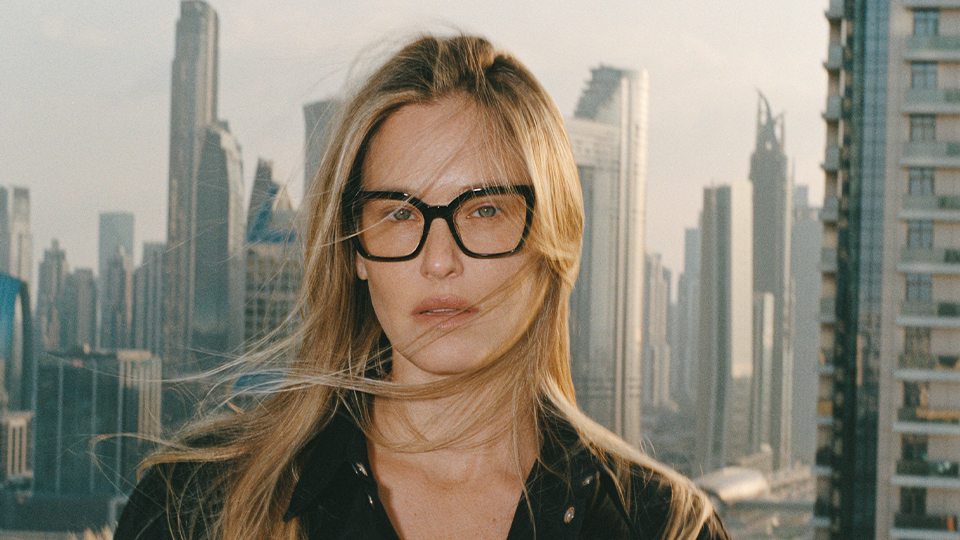 A model wears large square black spectacles and stands against a windy city background
